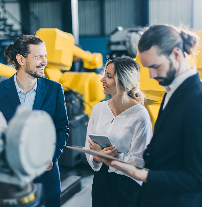A customer and two sales reps, in a discussion, standing in front of yellow industrial machinery, with one person holding a tablet